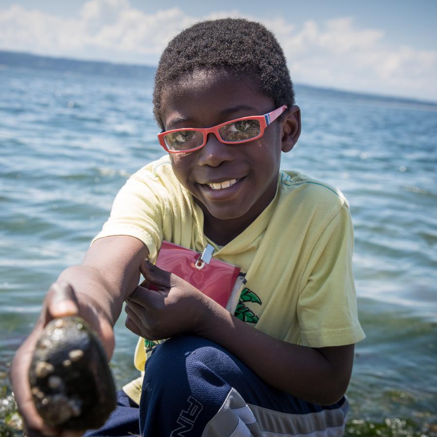 Smiling, black youth with a green shirt, blue pants, and red glasses displaying a rock they found on the beach. The rocky beach and Saratoga Pass are visible in the photo but are blurred.